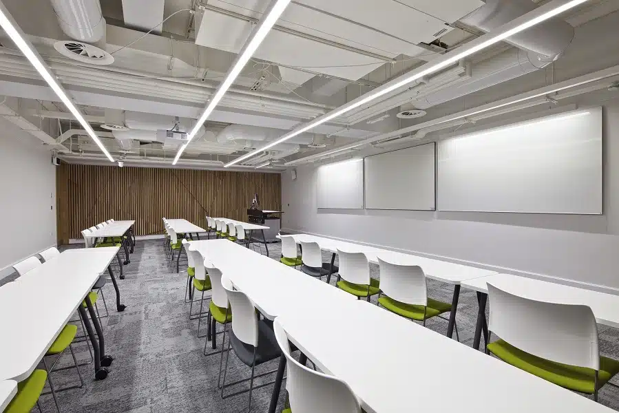 A small lecture theatre at UCL Torrington. There are white desks, chairs and boards. Some of the seats have a green cushion on them, whereas others have a grey one.