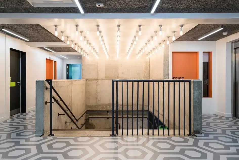 The top of a stairway in Notting Hill Prep School, there are various rows of bright lights on the ceiling, black railings and a patterned floor design at the top of the stairs.