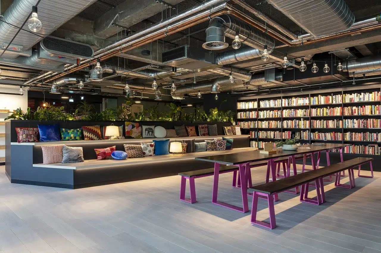 A coworking space that has a range of seating with various different cushions available. There is a black and pink working bench as well as a large bookcase in the background.