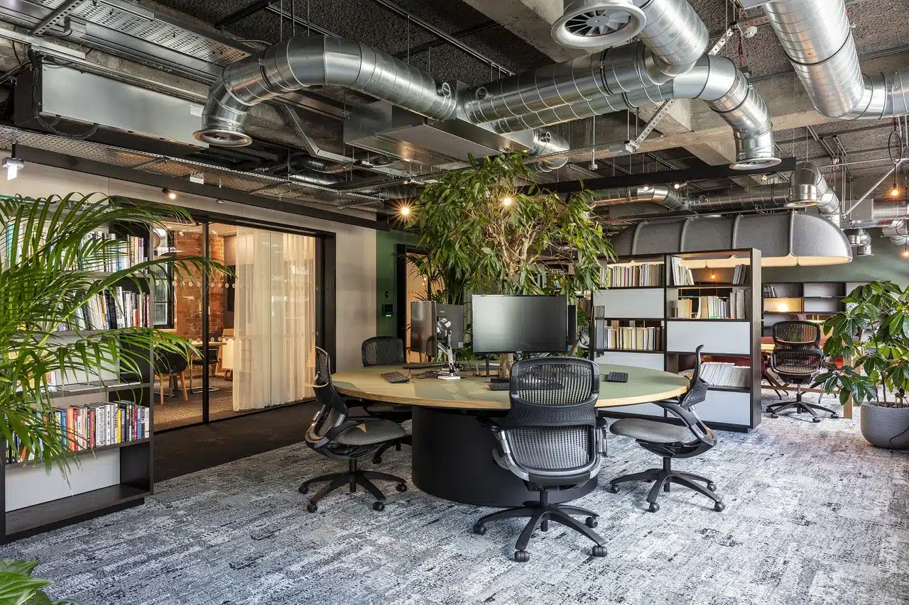 A round office table with various monitors and keyboards set up. There is a large tree coming up in the middle of the table. Office chairs surround the table and the carpet is grey.