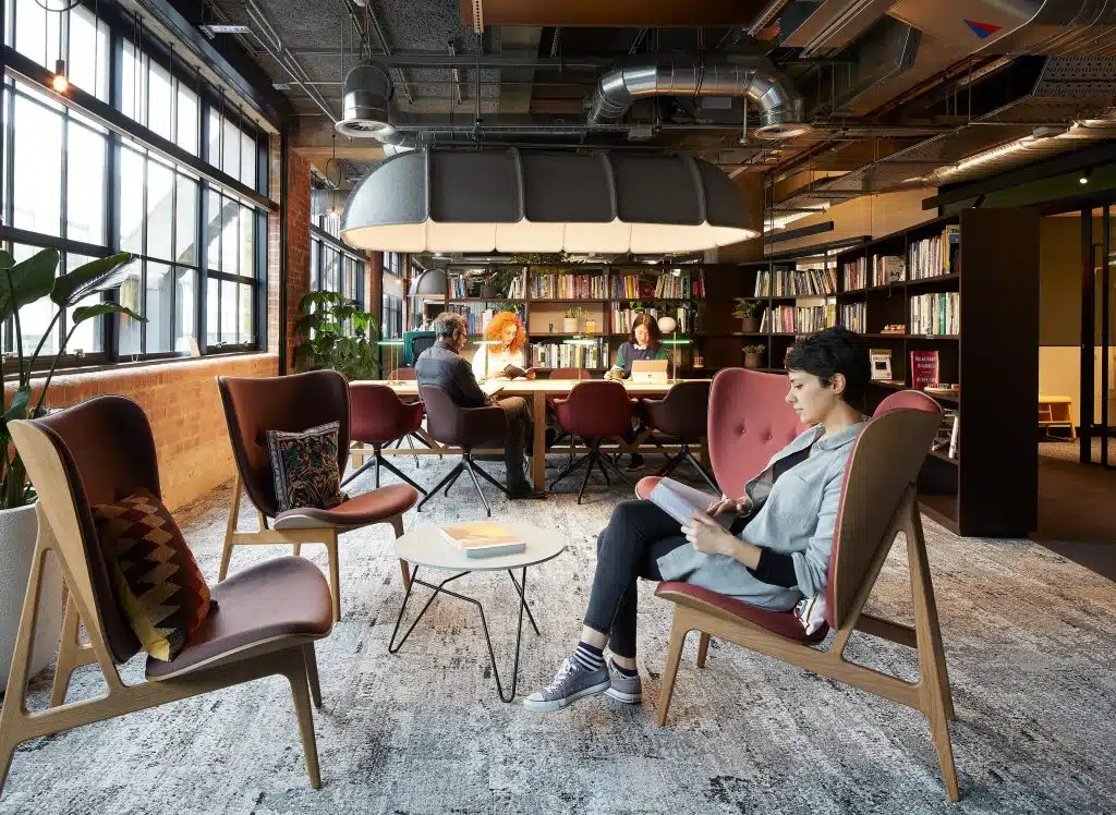 lady reading in chair in open plan offices with large windows and colleagues working behind her.
