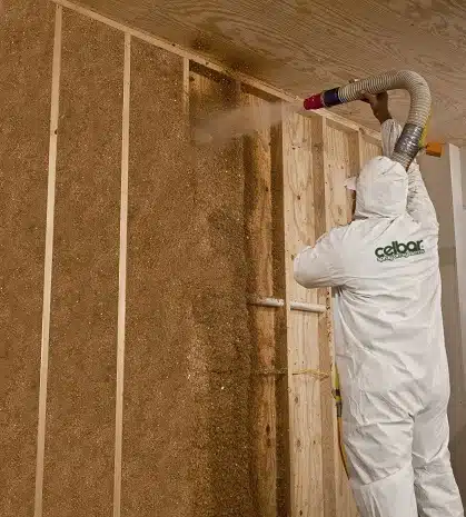 A worker in a protective outfit applying Celbar recycled insulation to a wooden wall in a building.