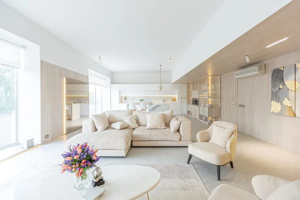 The interior of a living room that has white and beige furnishings. In the middle of the photo there is a beige corner sofa, just behind a small table and two armchairs. In the background there are two bar stools at a breakfast bar.