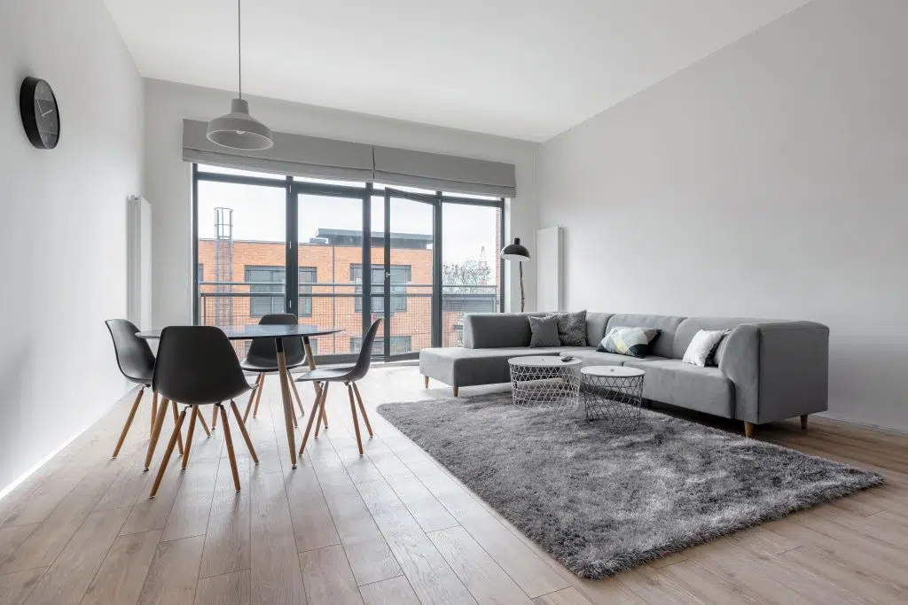 A grey corner sofa in a flat that has a kitchen table and two side tables next to it, and is located in front of some open doors which open onto a balcony. A floor lamp is overlooking the sofa. The room is finished with an acoustic ceiling.
