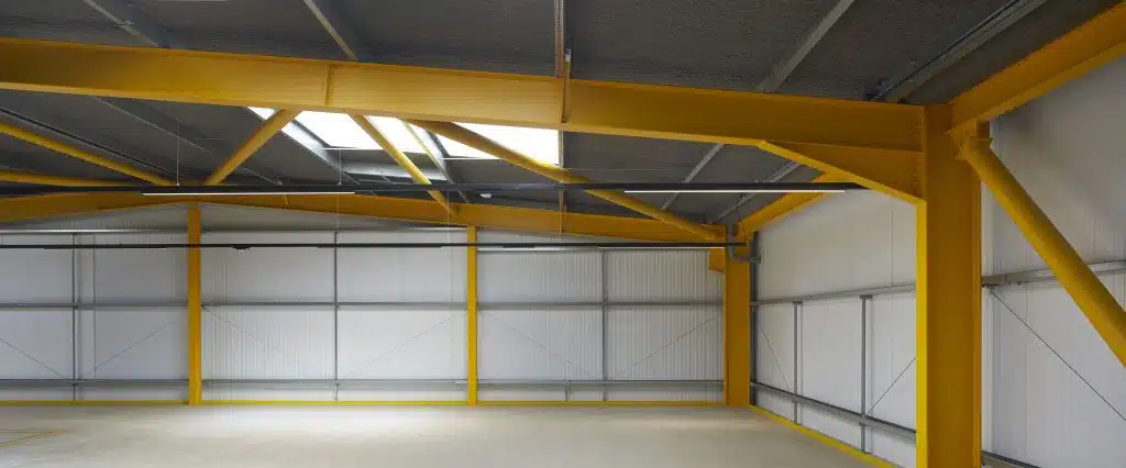 An interior view of an empty industrial warehouse showcasing yellow steel beams and grey walls. Bright natural light floods the space through skylights on the ceiling.