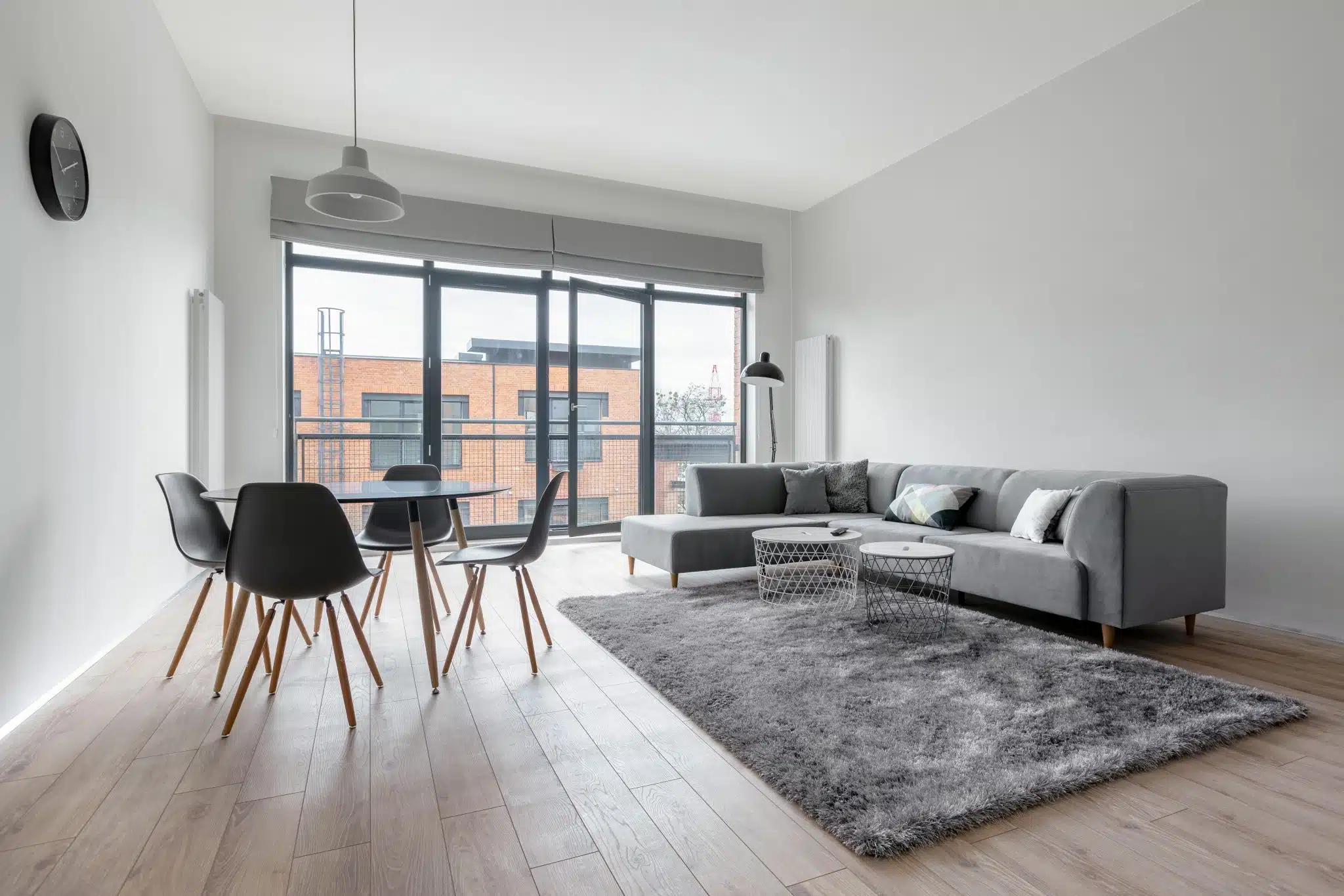 A grey corner sofa in a flat that has a kitchen table and two side tables next to it, and is located in front of some open doors which open onto a balcony. A floor lamp is overlooking the sofa.
