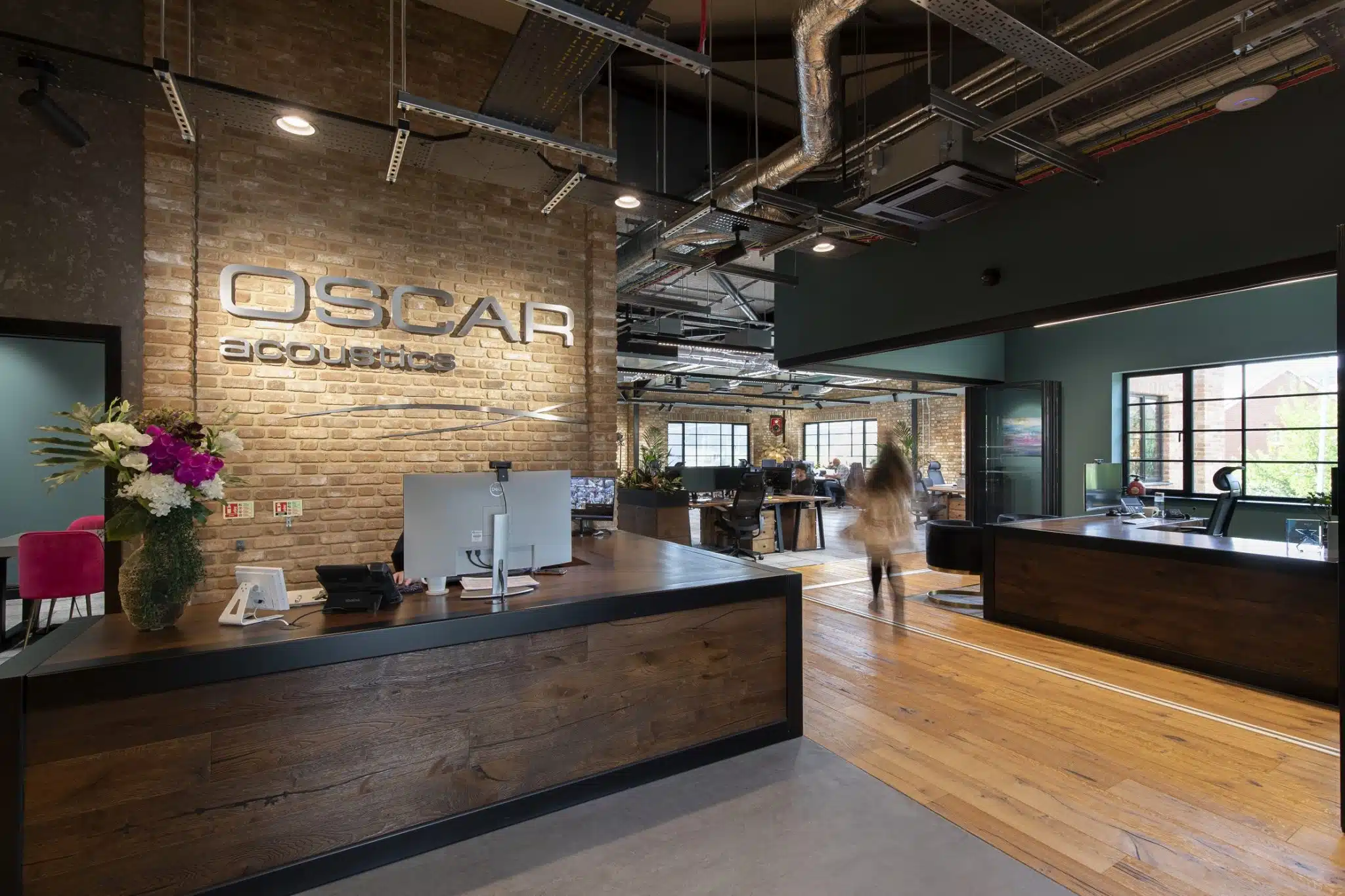 a wooden reception desk in front of a brick interior wall with 'Oscar Acoustics' signage. Behind are open plan offices with a figure walking across the wooden floor.