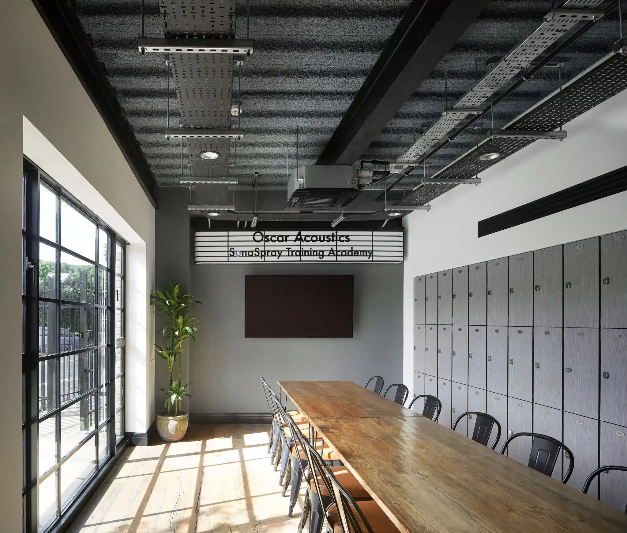 Oscar Acoustics' SonaSpray Training Academy. Grey lockers, TV, long wooden table and plants and windows.