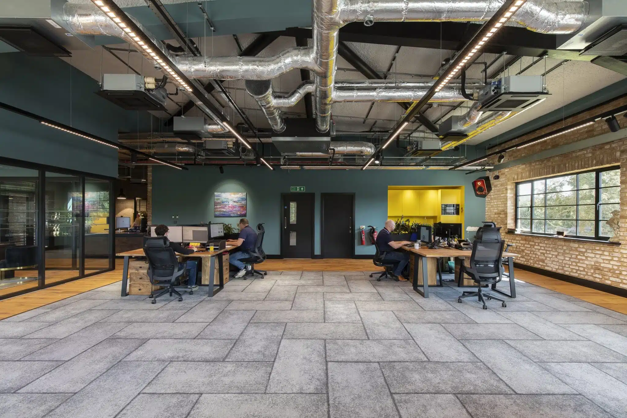 Open plan office with visible services on the ceiling and a grey acoustic spray for industrial sprayed concrete look. 2 men working at their desks and a bright yellow kitchen area behind.